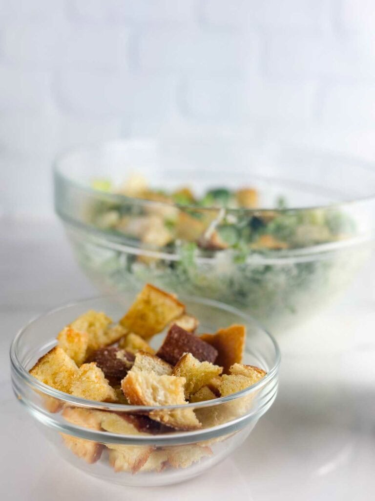 Homemade croutons in a clear bowl in front of a caesar salad.