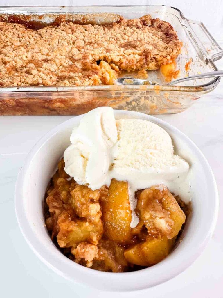 Serving of peach cobbler with vanilla ice cream in front of a baking dish of it.