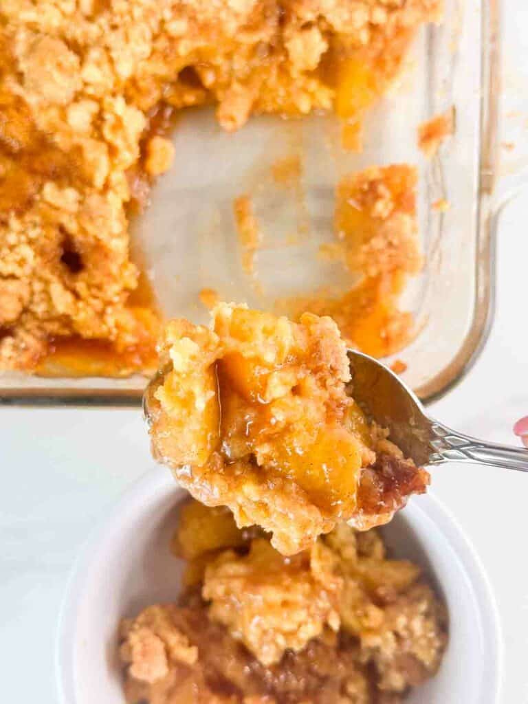 Spoon holding peach cobbler with cake mix over a bowl of it.
