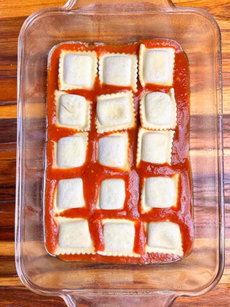 Ravioli on top of sauce in a clear baking dish.