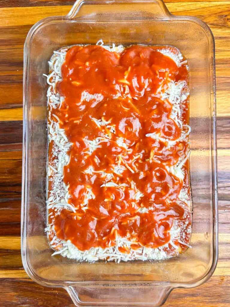 Some red sauce on top of cheese in a clear baking dish.