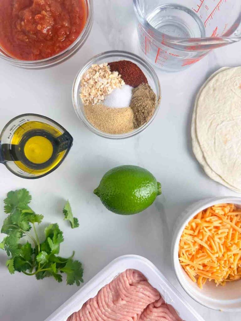 Ground chicken taco meat ingredients: ground chicken, lime juice, olive oil, chili powder, cumin, garlic powder, salt, dried onion, and water. Other ingredients for tacos: salsa, tortillas, cheese, and cilantro.