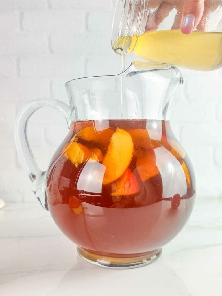 Pouring peach simple syrup into a pitcher of tea with peaches floating in it.