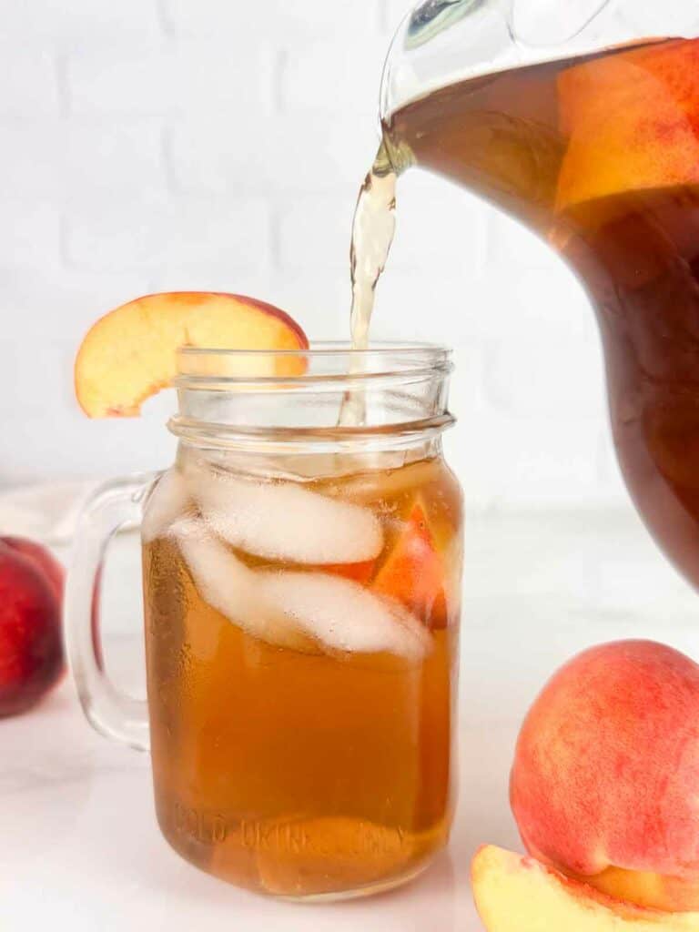 Pouring peach tea into a mason jar with ice and peach slices.