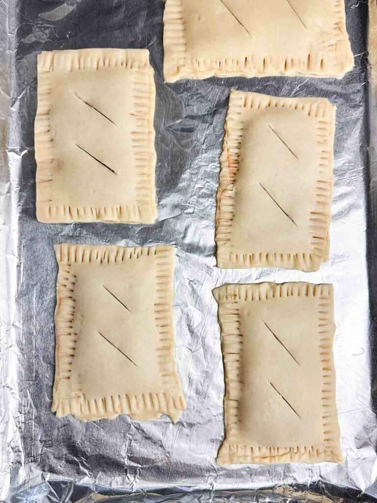 5 unbaked homemade strawberry pop tarts on a baking tray with crimped edges and 2 vents cut in the middles of each pop tart