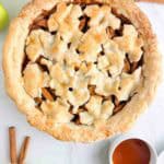 Apple pie with leaf design on top next to caramel sauce and cinnamon sticks.