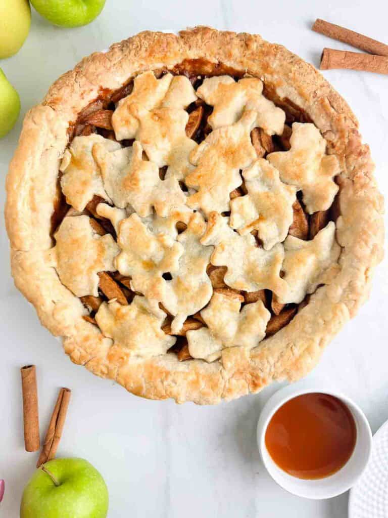 Apple pie with leaf design on top next to caramel sauce and cinnamon sticks.