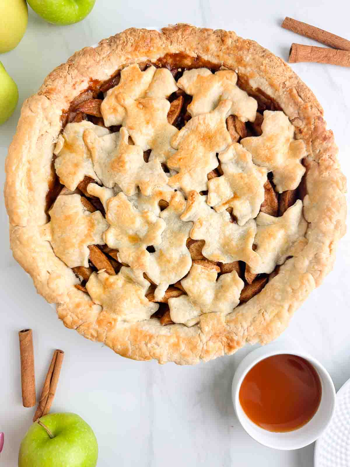 Apple pie with leaf design on top next to caramel sauce and cinnamon sticks.