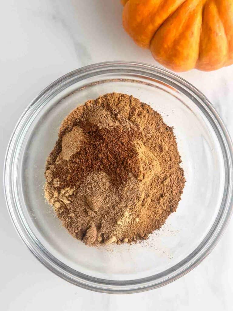 Spices for pumpkin pie spice recipe in a small clear bowl with a pumpkin next to it.