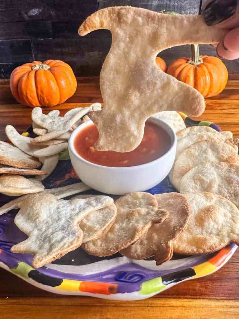 A plate of Halloween tortilla chips shaped like black cats, witch hats, and pumpkins with a black cat chip ready to be dipped in salsa