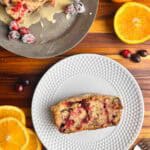 Cranberry orange bread with walnuts on a plate and on a serving tray with cranberries and oranges surrounding them.