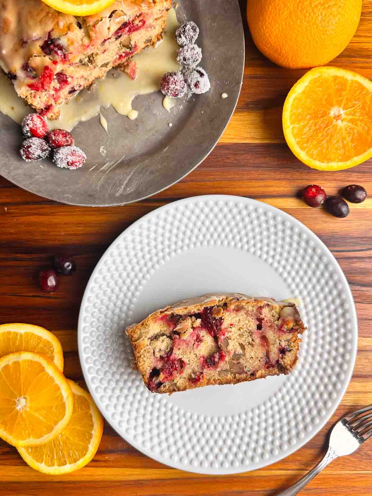 Cranberry orange bread with walnuts on a plate and on a serving tray with cranberries and oranges surrounding them.