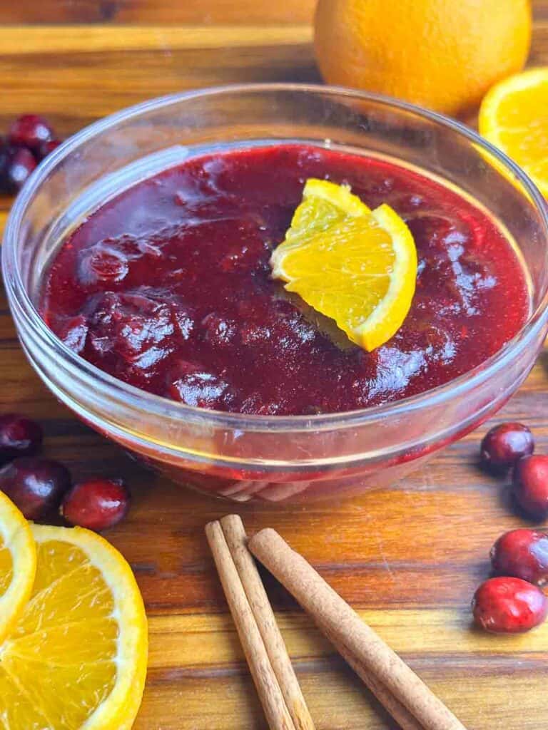 Orange cranberry sauce in a clear bowl with orange slices, cinnamon sticks, and cranberries.