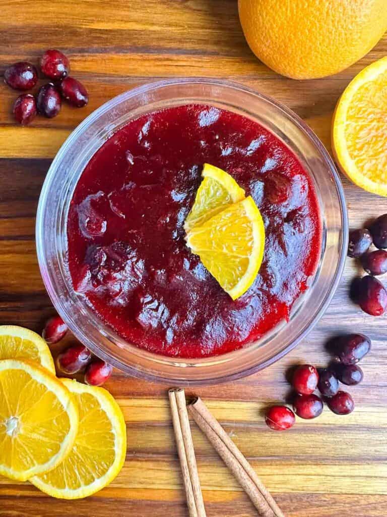 Orange cranberry sauce in a clear bowl with orange slices, cinnamon sticks, and cranberries.