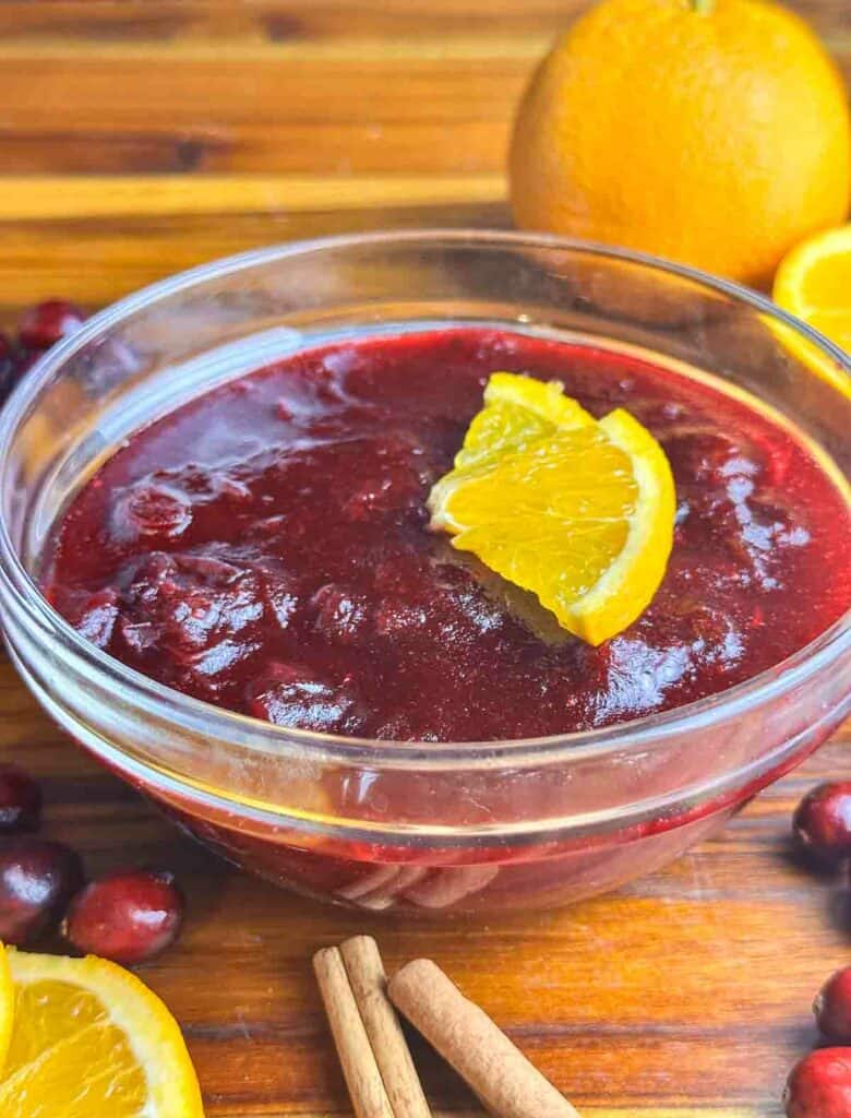 Orange cranberry sauce in a clear bowl with orange slices, cinnamon sticks, and cranberries.
