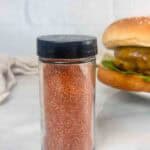Jar of homemade burger seasoning with a burger in the back made with it.