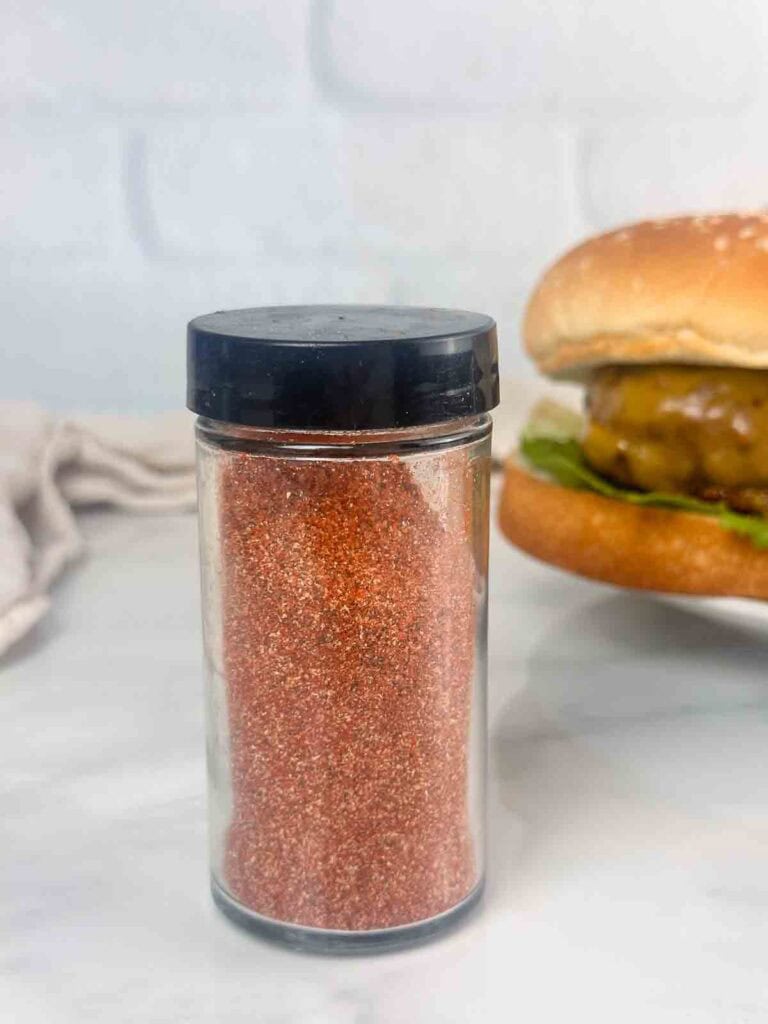 Jar of homemade burger seasoning with a burger in the back made with it.