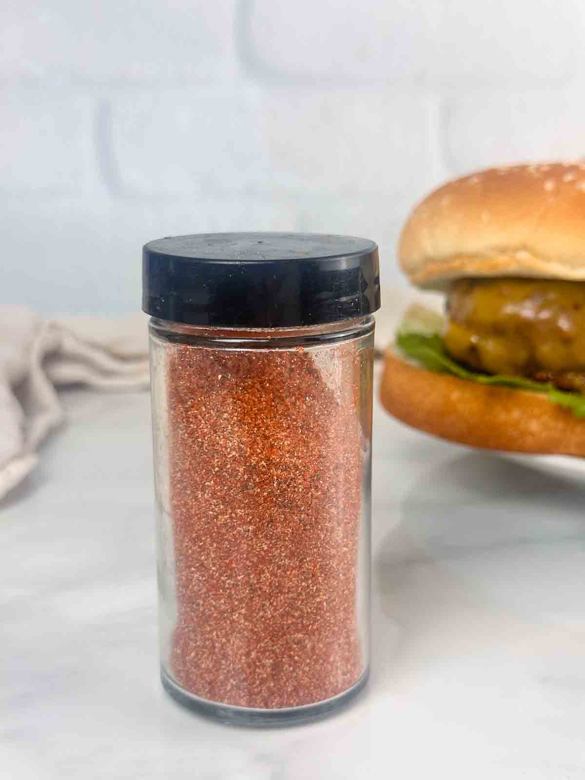 Jar of homemade burger seasoning with a burger in the back made with it.