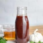 A bottle of homemade honey barbecue sauce on a counter with a jar of honey and head of garlic
