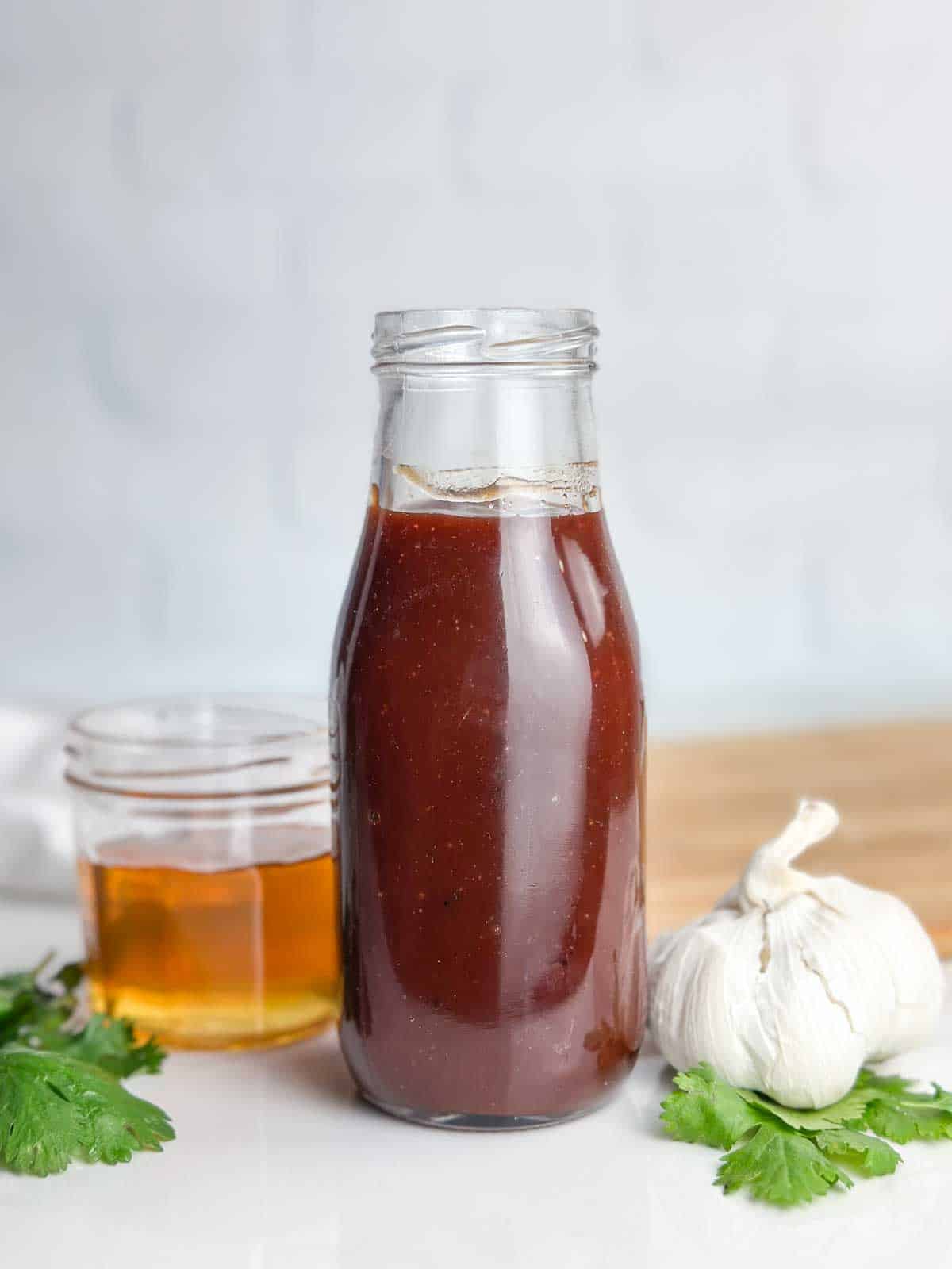 A bottle of homemade honey barbecue sauce on a counter with a jar of honey and head of garlic